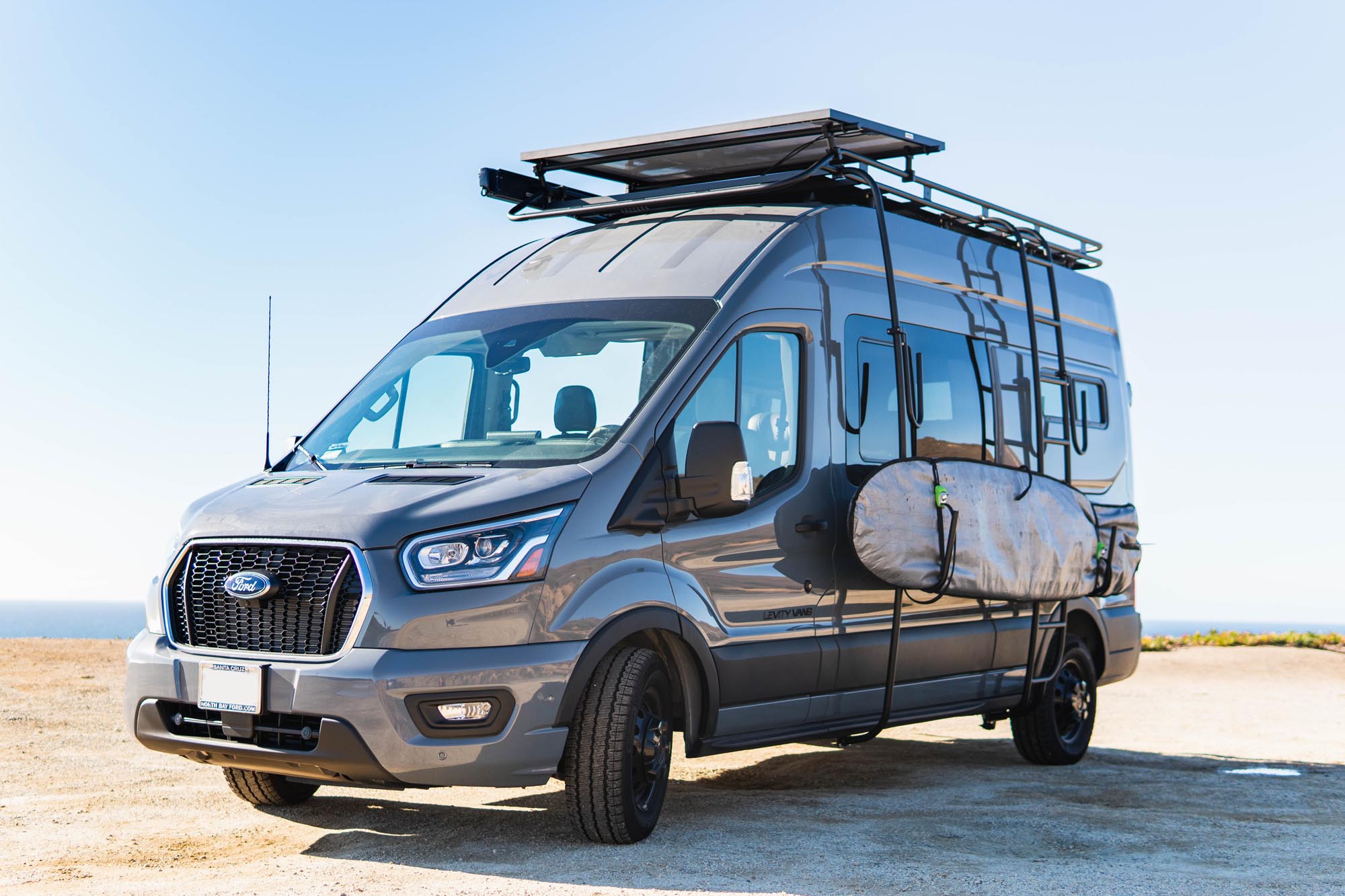 ford transit van conversion with external surfboard rack, roof rack, and solar panel parked on a beach near the ocean